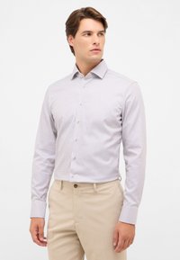 Hemd mit Knopfleiste aus leichtem Stoff mit kleinem blauen Muster, spitzem Kragen und langen Ärmeln. Kombiniert mit beigen Hosen.