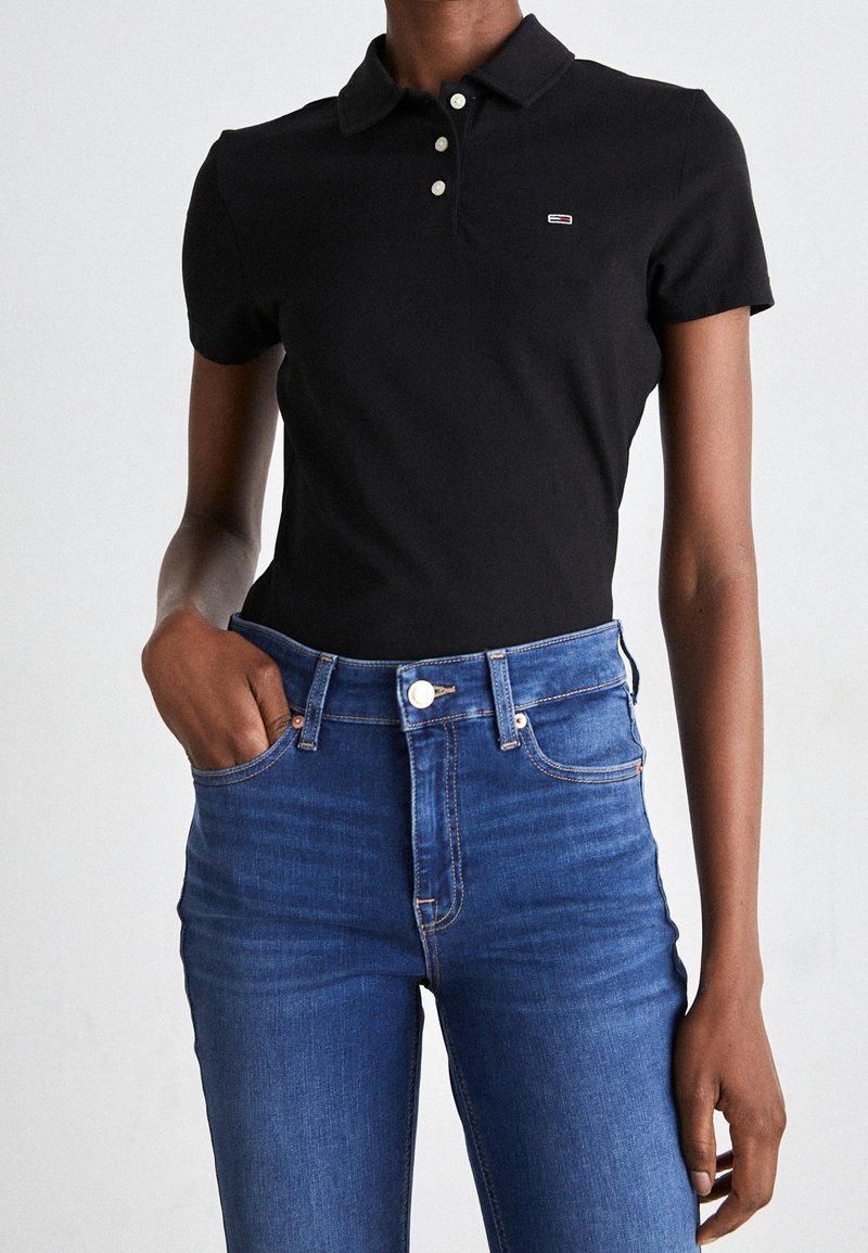 Personne portant un polo noir à manches courtes rentré dans un jean bleu taille haute, une main dans la poche.