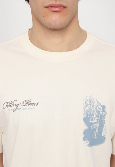 T-shirt en coton crème avec un graphique bleu représentant une personne s'occupant des plantes et un texte brodé "Filling Pieces" avec un slogan.