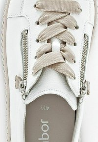 Baskets en cuir blanc avec des lacets beige clair, deux zips sur les côtés et une surface texturée. La semelle intérieure est grise avec un logo.