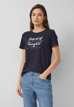 Frau mit braunen Haaren, die ein dunkelblaues T-Shirt mit motivierendem Text und hellblaue Jeans trägt, steht vor einem einfarbigen grauen Hintergrund.