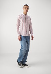 Homme portant une chemise à rayures rose et blanche à manches longues, un jean bleu clair ample et des baskets noires, se tenant sur un fond blanc uni.