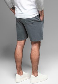Pantalones cortos de algodón gris con corte recto, que cuentan con bolsillos laterales y un bolsillo trasero, combinados con zapatillas blancas, mostrados desde la parte trasera.
