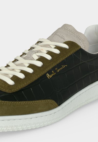 Baskets basses en daim olive, panneaux en cuir noir à texture crocodile, lacets blancs, semelle blanche, et logo doré "Paul Smith" sur le côté.