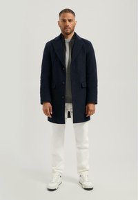 Marine blauwe wollen jas met een notch-kraag, enkelvoudige sluiting, zijzakken, gedragen over een grijze trui en witte broek, gecombineerd met witte sneakers.