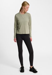 Langärmliges, hellgrünes Sporttop, kombiniert mit schwarzen Leggings und schwarzen Sneakers. Das Top hat einen runden Halsausschnitt und eine entspannte Passform.