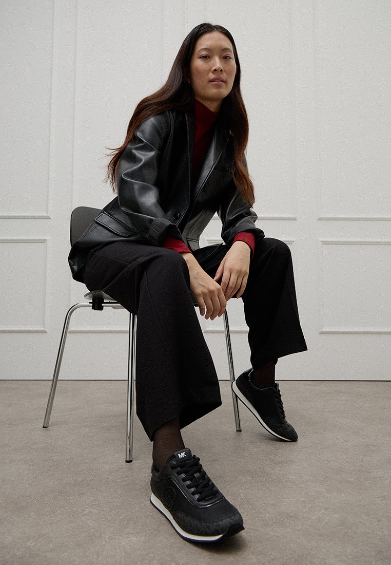 Vrouw met lang zwart haar, gekleed in een zwart leren jack, bordeauxrode coltrui, zwarte wijde broek en zwarte sneakers, zittend op een stoel.