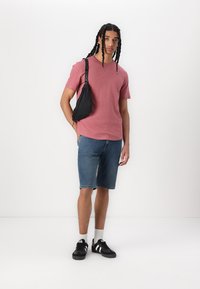 Rdeča bombažna majica, kavbojke kratke hlače, črna torbica za čez ramo, črni športni copati z belimi črtami, stoji na svetli podlagi.