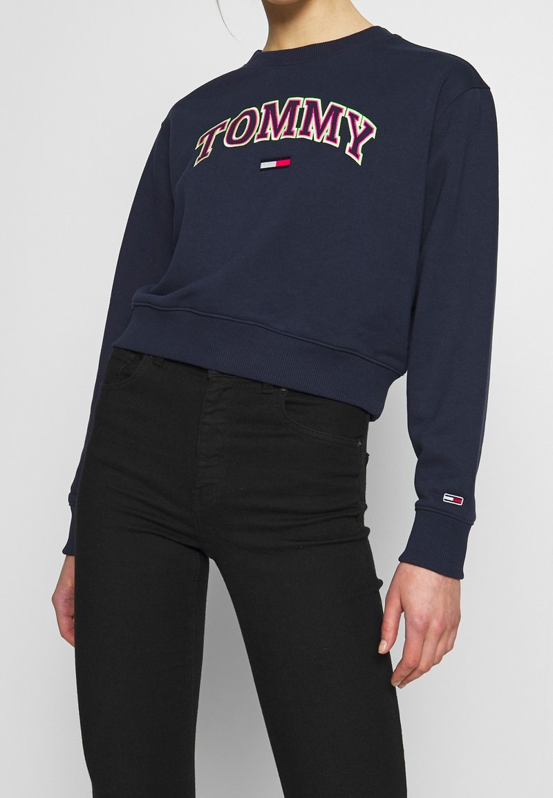 Personne portant un sweat-shirt bleu marine avec "TOMMY" en grandes lettres et un logo sur la manche, associé à un jean noir taille haute.