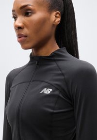 Μαύρο αθλητικό μπουφάν με φερμουάρ, κατασκευασμένο από λεία, ελαστική ύφανση. Διαθέτει ανακλαστικό λογότυπο στο στήθος και ψηλό γιακά.