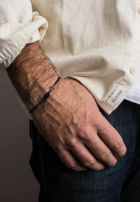 Brățară împletită neagră cu accente metalice, având un șarm rotund și detalii cu lanț, purtată pe o încheietură împotriva unei cămăși de culoare deschisă.