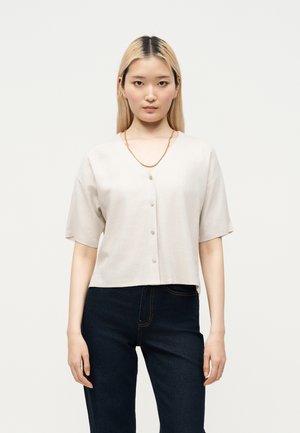 Jeune femme aux cheveux blonds raides portant une chemise beige à manches courtes boutonnée et un jean foncé, debout devant un fond blanc uni.