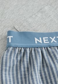 Material gri cu un tiv elastic albastru, având textul alb "NEXT". Partea inferioară are dungi alternative albastre și albe.