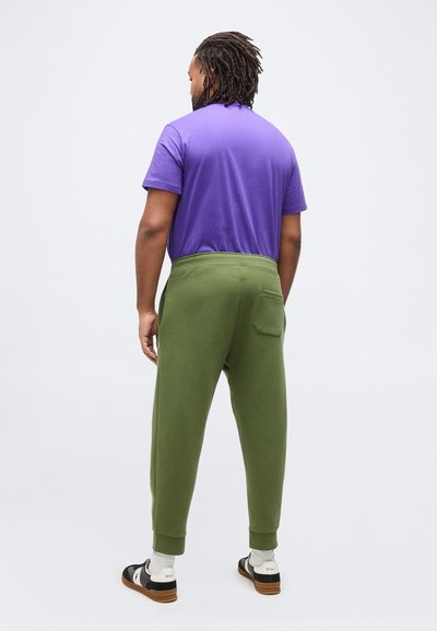 Άντρας με dreadlocks στέκεται γυρισμένος με την πλάτη φορώντας μωβ πουκάμισο, χακί παντελόνι jogging, λευκές κάλτσες και μαύρα και λευκά αθλητικά παπούτσια.