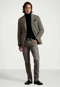 Moški nosi sivi blazer, črn pulover z visokim ovratnikom, sive kavbojke in črne mokasine, stoji z eno roko v žepu.