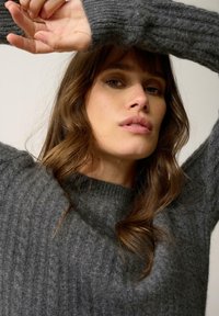 Szary sweter z prążkowanej dzianiny o okrągłym dekolcie. Materiał wydaje się miękki i teksturowany, ma długie rękawy oraz subtelne różnice w kolorze.