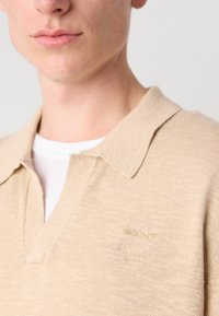 Polo din tricot bej cu o textură moale și un guler. Prezintă un logo subtil în aur pe partea stângă. Este purtat peste o cămașă albă.