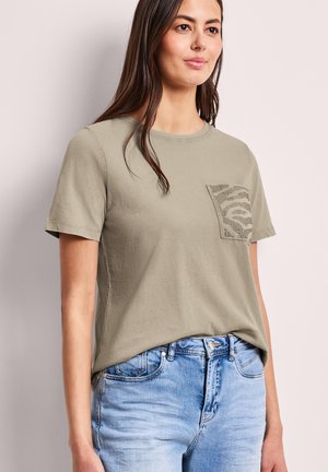 Donna che indossa una t-shirt a maniche corte verde oliva chiaro con tasca a stampa zebrata testurizzata e jeans azzurri, in piedi contro uno sfondo chiaro.