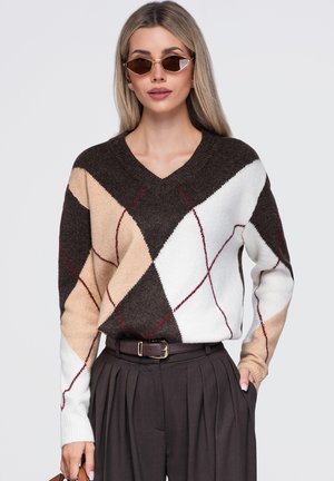Femme portant un pull à losanges marron, beige et blanc avec un pantalon foncé et des lunettes de soleil œil de chat sur un fond uni.