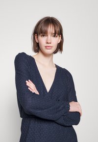 Vestido azul-marinho com decote em V profundo, mangas longas com punhos elásticos, tecido texturizado e cintura ajustada, apresentando um padrão de diamante subtil.