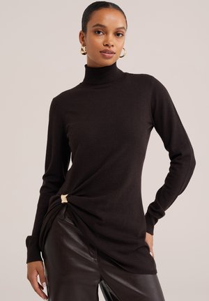 Femme portant un pull à col roulé noir avec un détail de fermoir doré et un pantalon en cuir foncé, posant avec une main sur la hanche.