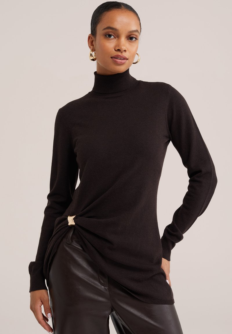 Femme portant un pull à col roulé noir avec un détail de fermoir doré et un pantalon en cuir foncé, posant avec une main sur la hanche.