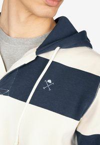 Sudadera con capucha que presenta rayas horizontales en tonos azul marino y crema, detalles de costura y un logo con iconos cruzados en el pecho.