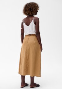 Witte, mouwloze top met een V-hals en strikbandjes; mosterdkleurige rok tot halverwege de kuit met een zijrits en subtiele textuuraccenten; zwarte sandalen.