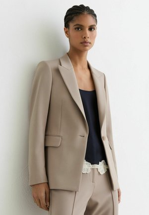 Kvinde med flettet hår iført en beige blazer og bukser, sort top med hvid blonderkant, står op ad en hvid væg og kigger fremad.