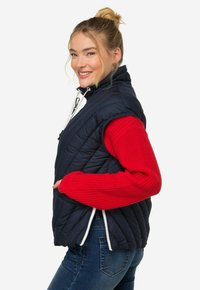 Marineblå puffväst med hög krage, vit dragkedja och quiltat mönster, ihop med en röd stickad tröja. Modellen poserar med ett leende.