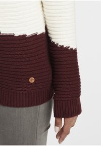 Pull en maille avec rayures horizontales bordeaux et crème, texture côtelée et un logo décoratif en bois sur l'ourlet inférieur.