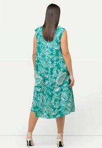 Kvinna med rakt brunt hår bär en ärmlös turkos klänning med vita lövmönster, står vänd med ryggen mot kameran, vita högklackade sandaler.