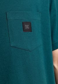 Tyrkysové bavlněné tričko s náprsní kapsou a černou logo nášivkou. Měkká textura, kulatý výstřih a krátké rukávy.