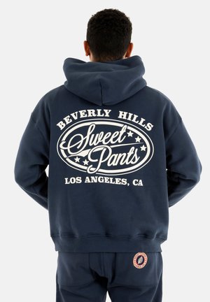 Sudadera azul marino con un gran gráfico blanco en la parte trasera que dice "Sweet Pants" y "Beverly Hills, LOS ÁNGELES, CA." Presenta un bolsillo canguro.