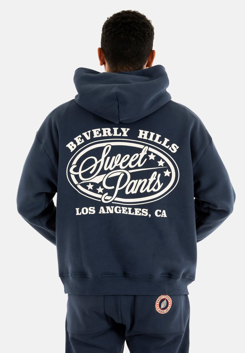 Hoodie bleu marine avec un grand graphique blanc au dos portant les inscriptions "Sweet Pants" et "Beverly Hills, LOS ANGELES, CA." Il dispose d'une poche kangourou.
