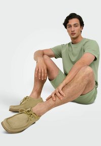 Lys grøn, struktureret kortærmet shirt og shorts, parret med beige ruskindssko med snørebånd og flad sål, siddende på en neutral baggrund.