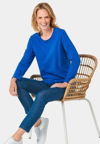 Blauw langmouwen shirt van zacht materiaal, ronde hals, gecombineerd met donkere denim jeans en witte sneakers, zittend op een gevlochten stoel.