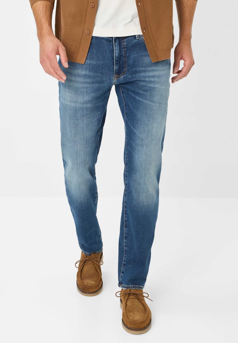 Jeans in denim azzurro chiaro con un effetto leggermente sbiadito, taglio dritto e cuciture gialle discrete, abbinati a scarpe in suede marroni.