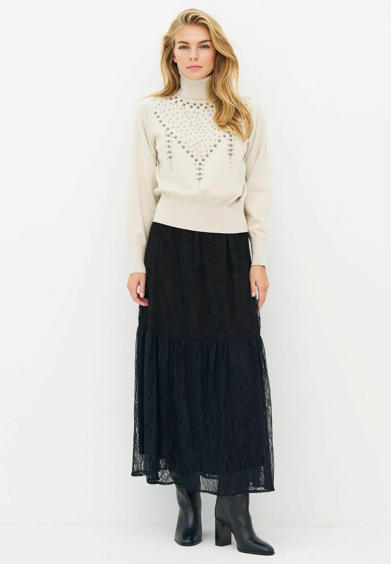 Femme aux longs cheveux blonds, portant un pull à col roulé beige clouté, une jupe midi en dentelle noire et des bottines noires à talons, debout sur fond blanc.
