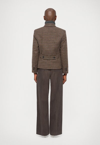 Hnedý houndstooth sako s priliehajúcim pásom, dvojitými gombíkmi a šedým vysokým golierom, skombinované s tmavými, širokými rebrovanými nohavicami.