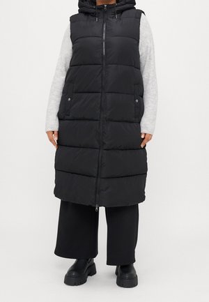 Person iført en lang sort polstret vest med hætte, grå langærmet top, brede sorte bukser og sorte kraftige støvler, stående på en hvid baggrund.