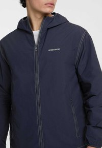 Giacca con cappuccio di colore navy realizzata in tessuto morbido, con cuciture bianche e una zip frontale. Logo "Guess Jeans" sul lato sinistro del petto, tasche laterali.