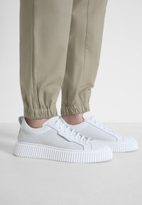 Witte sneakers met een gestructureerde bovenkant, voorzien van geperforeerde details en een gegroefde zool. Gdragend met beige opgekrulde broeken.