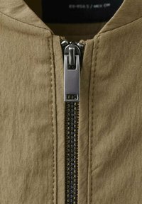 Detailný záber na khaki látku s metalickým zipsom so štvorcovým ťahákom, zvýrazňujúci textúrovaný povrch a detailné prešitie vedľa zipsu.