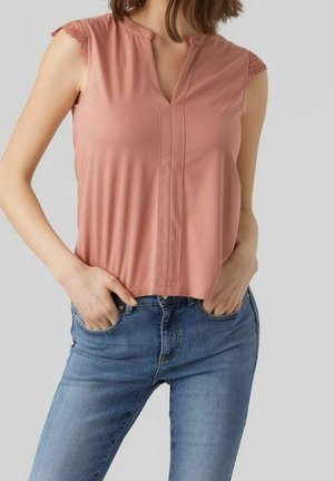 Frau trägt ein ärmelloses, staubrosa Oberteil mit Spitzenbesatz und blaue Jeans, steht mit den Händen in den Taschen vor einem schlichten Hintergrund.