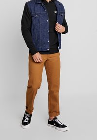 Gilet in denim con due tasche sul petto, indossato sopra un hoodie nero. Pantaloni marroni e sneakers nere con dettagli bianchi completano l'outfit.