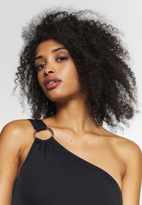 Maillot de bain noir à une épaule avec une bride froncée et un accent en anneau argenté. Sa texture lisse et sa coupe ajustée mettent en valeur la silhouette.