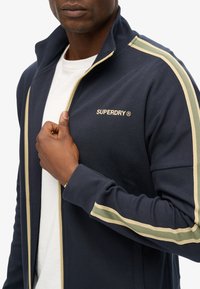 Veste zippée marine en mélange de coton avec des rayures vert clair et crème sur les manches, arborant un logo doré "SUPERDRY" sur la poitrine.
