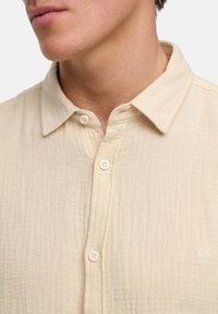 Beige Hemd mit Knöpfen aus strukturiertem Stoff, mit Kragen und drei Knöpfen. Enthält ein kleines gesticktes Logo auf der Brust.