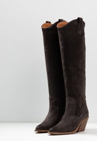 Un par de botas altas de ante marrón oscuro con puntas afiladas y tacones apilados de madera, de pie sobre una superficie blanca.